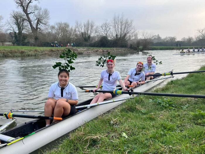 Smiling rowers sat in the boat by the grassy riverbank with greenery in their hair.