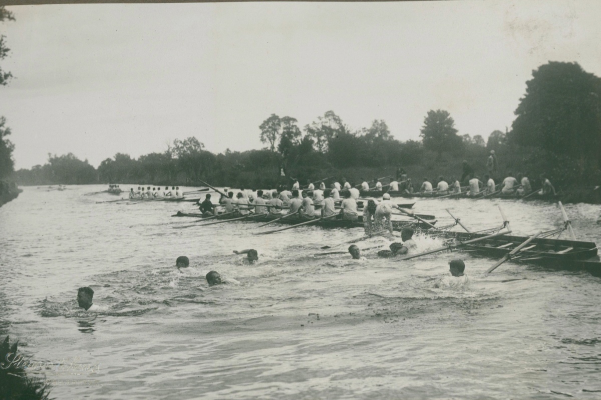 Rowers swimming in the river as boats continue to race in the background.