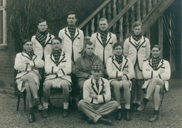 Students dressed in blazers and tartan scarves, posed in front of the Trinity Hall Boat House.