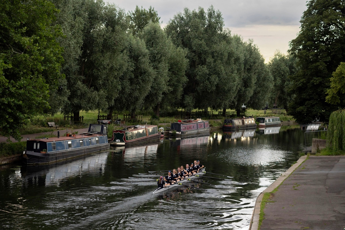 A rowing crew in matching dark kit rows along a calm river lined with narrowboats and tall trees. The boat moves through the water beneath an overcast sky, with reflections of the boats and trees visible on the river’s surface.