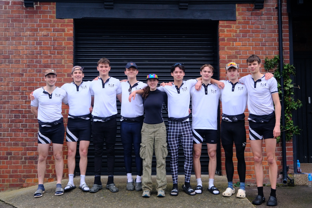 A rowing squad in matching black-and-white kit stands in a line in front of a black shutter and brick building. One person wearing a dark top and green trousers stands in the centre, with team members linking arms around each other for a group photograph.