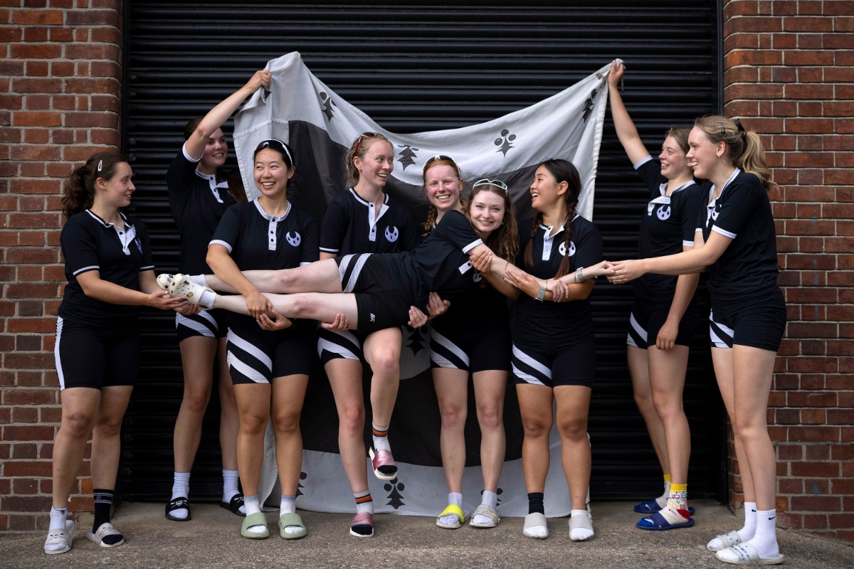 A rowing squad in matching black-and-white kit poses in front of a black shutter framed by brick walls. Several team members hold up a black-and-white flag behind the group while others lift a teammate horizontally across their arms. Everyone is wearing training clothing and sliders or socks, creating a relaxed team portrait.